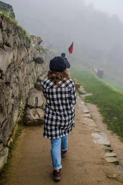 Woman walking along an ancient stone path in misty weather, dressed in a plaid shirt and hat. Ideal for themes related to adventure, travel, nature walks, history tours, and outdoor explorations. Useful for travel blogs, adventure tour promotions, and nature photography portfolios.