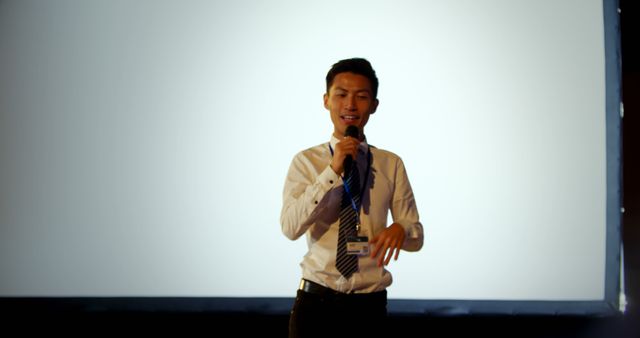 Young male professional, standing on stage, confidently presenting with a microphone. Useful for themes related to business seminars, leadership training, public speaking sessions, and corporate events. Suitable for illustrating concepts of confidence, professionalism, and effective communication.