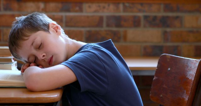 Young boy in a school setting, resting his head on his desk, likely exhausted or taking a nap. Ideal for illustrating concepts of tired children, the need for breaks during school hours, study fatigue, or educational environments. Useful for articles about childhood education, mental health in schools, or parent recommendations.
