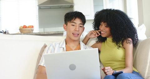 Multiracial couple sharing laptop on cozy living room sofa, collaborating and smiling