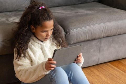 School-age girl sitting on hardwood floor holding tablet beside gray sofa, focused