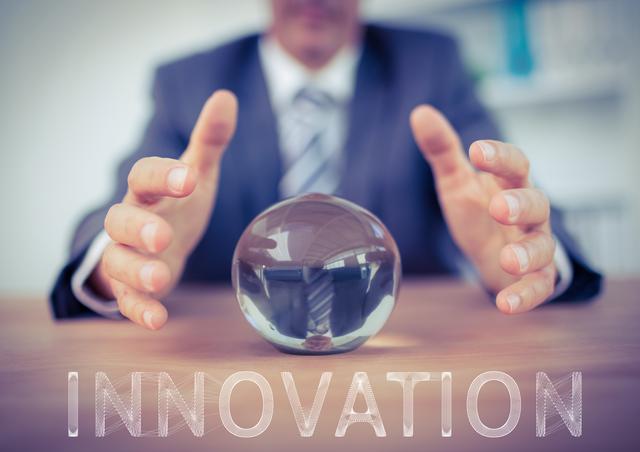 Businessman wearing a suit holding hands around a clear crystal ball with 'Innovation' text overlay on wooden table background. Could be used in contexts emphasizing futuristic business strategies, technology-driven approaches, corporate innovation management, strategic planning, and leadership envisioning. Suitable for websites, presentations, and articles focused on modern business practices and technological advancements.