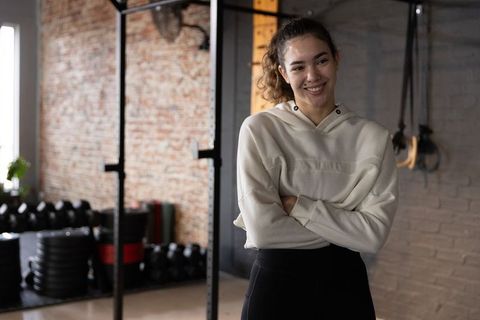 Smiling woman in hoodie with arms crossed beside squat rack and weights, gym confidence