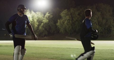 Night cricket practice: two batsmen walking across floodlit outfield carrying bats and helmets
