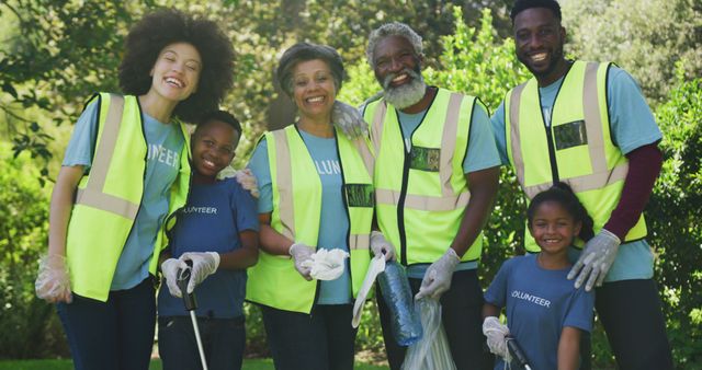 Diverse group of volunteers wearing safety vests cleaning park while smiling for the camera. Great for promoting community service initiatives, environmental campaigns, and teamwork. Also useful for illustrating concepts of family bonding and volunteer work in diverse communities.