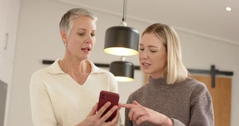 Mature and middle-aged women comparing smartphone and pointing while browsing in modern kitchen