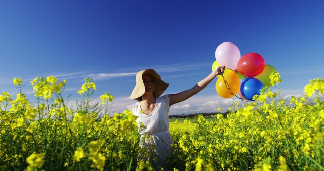 A young Caucasian girl enjoys a sunny day in a vibrant yellow flower field, holding colorful balloons, with copy space. Her wide-brimmed hat and bright balloons add a playful touch to the idyllic rural landscape.