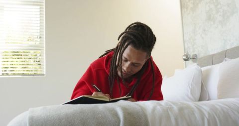 African american man writing in journal on bed wearing red hoodie relaxed modern bedroom