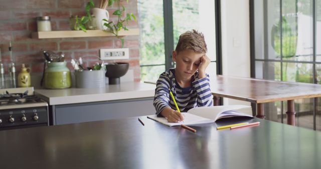 Young boy sitting at kitchen table studying and writing in a book with pencils scattered nearby, in a bright and modern kitchen with an open layout and natural light. Can be used in educational materials, parenting blogs, home schooling resources, and advertisements for tutoring services or home decor.