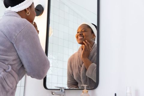 Smiling african american woman grooming face at bathroom mirror wearing robe and headband