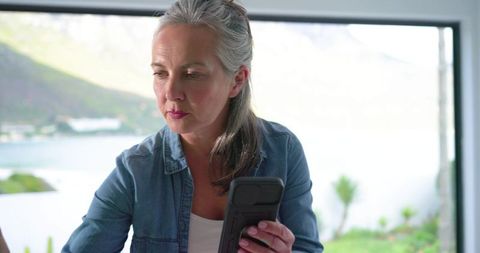 Mature woman holding smartphone gazing thoughtfully through window with lakeside view