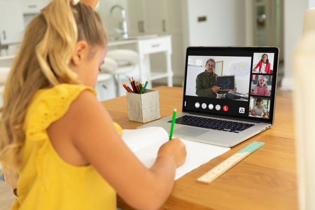 Caucasian girl with blond hair writing notes while learning online over video call on laptop. Home, screen, unaltered, childhood, wireless technology, education, student and e-learning concept.