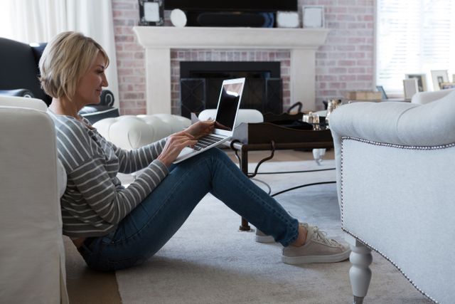 Woman sitting on the floor in a modern living room using a laptop. Ideal for illustrating remote work, home office, lifestyle, and technology in a cozy home environment. Suitable for articles on home-based productivity, casual work settings, or modern living spaces.