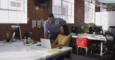 Woman typing on laptop with colleague leaning over in bright loft office with exposed brick walls