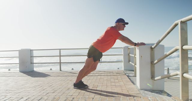 Man in hat and sunglasses is performing push-ups against a railing by the sea on a sunny day. The setting includes a boardwalk with ocean waves in the background. Ideal for use in health and fitness articles, promotional materials for athletic apparel, or content emphasizing outdoor exercise.