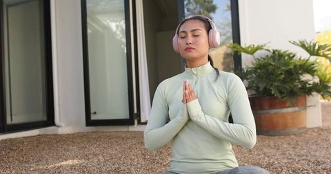 Mid-adult asian woman meditating with over-ear headphones on modern patio in athletic wear