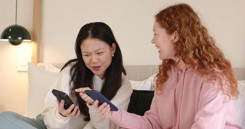 Asian and redhead friends sharing smartphones on cozy bed, laughing and comparing screens