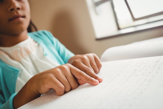 Young girl reading a braille book in a classroom. Ideal for educational materials, articles on accessibility and inclusion, special education resources, and promoting literacy for visually impaired students.