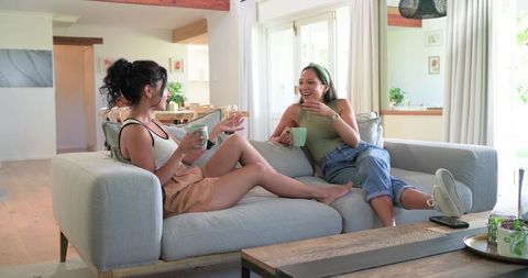Indian and african american friends laughing on sofa, coffee chat in sunlit living room