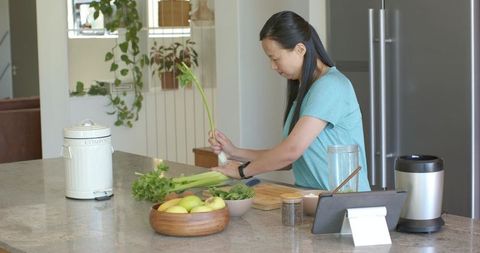 Asian woman preparing celery on modern kitchen island with apples, compost bin, smartwatch