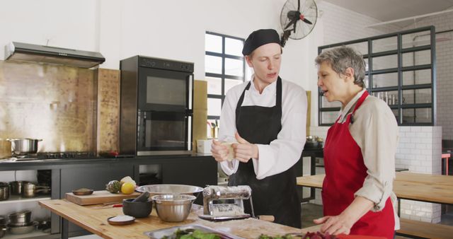 Professional chef instructing senior woman in modern kitchen environment. Senior woman wearing red apron, both engaging in food preparation activities. Ideal for use in content related to culinary classes, professional training, senior education, food preparation tutorials, and showcasing teamwork in kitchen settings.