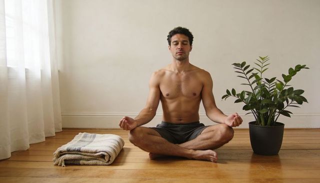 Shirtless man meditating cross-legged on hardwood floor with potted plant and blanket