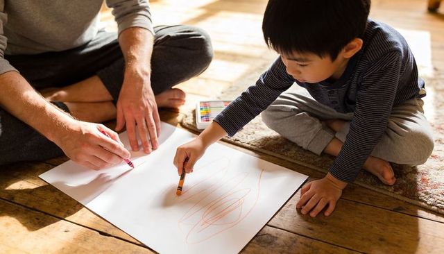 Father and son drawing together on sunlit wooden floor with crayons, creative bonding and learning