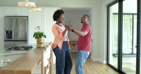 African American couple dancing and laughing in sunlit modern kitchen by wooden island