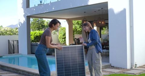 Multiracial women inspecting solar panel by backyard pool promoting home renewable energy