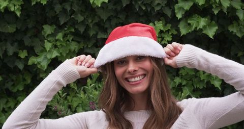 Cheerful woman adjusting santa hat amid ivy backdrop