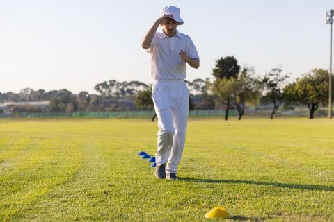 Male cricketer practicing cone agility drills at sunset wearing white bucket hat and whites