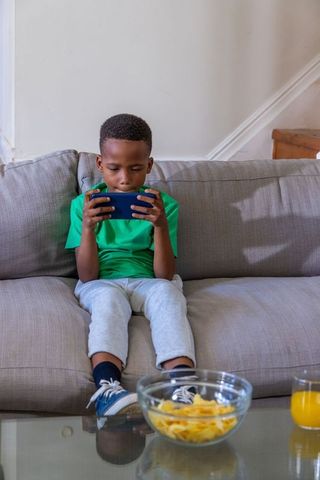African american boy playing with smartphone on couch at home with snacks and juice