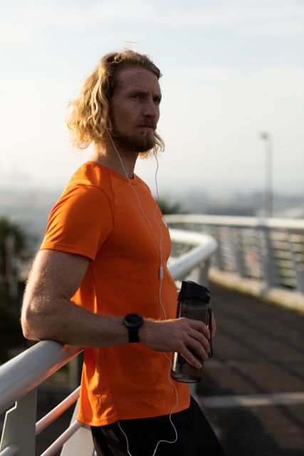 Side view of a fit Caucasian man with long blonde hair wearing sportswear exercising outdoors in the city on a sunny day with blue sky, standing and resting on a footbridge, holding water bottle in hand, wearing headphones.