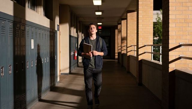 Male student walking down sunlit school corridor carrying notebooks and backpack by lockers