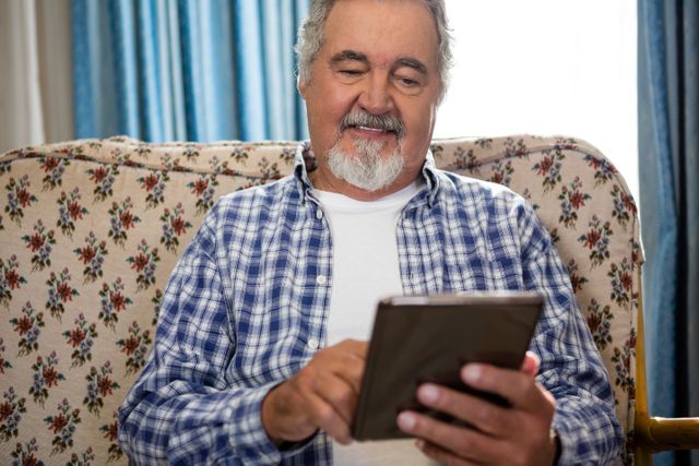 Senior man sitting on a floral-patterned sofa, using a digital tablet with a smile. He is wearing a plaid shirt and appears relaxed and comfortable. Ideal for use in articles or advertisements related to elderly care, technology for seniors, retirement living, and modern lifestyles for older adults.