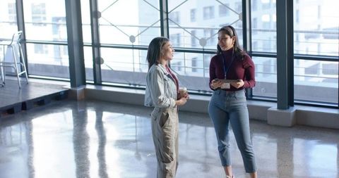 Diverse female colleagues chatting in modern atrium holding tablet and coffee cup