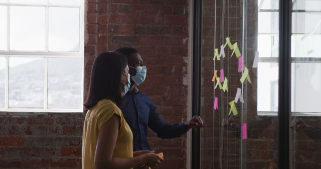 Colleagues wearing masks brainstorming ideas using colorful sticky notes on an office glass wall. Ideal for themes related to workplace safety during the pandemic, effective teamwork, collaboration, or brainstorming sessions.