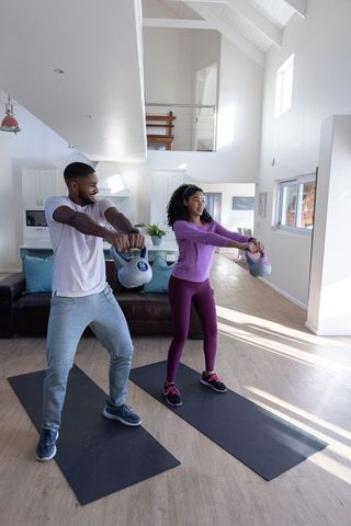 Diverse couple exercising with kettlebells in modern living room