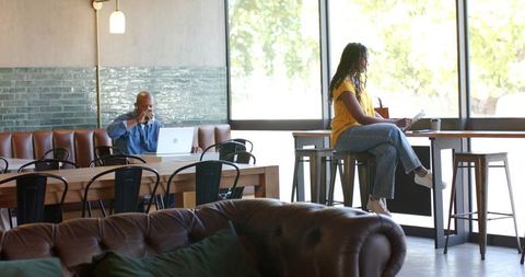 Sunlit cafe coworking: woman in yellow top working on laptop, sipping coffee by window