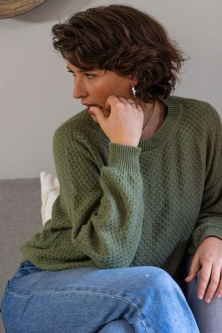 Young woman sitting on sofa wearing olive-green sweater and jeans looking contemplative