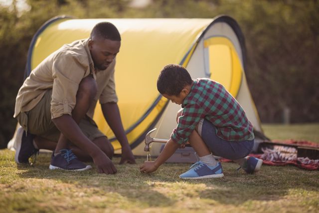 Father and son working together to set up a yellow tent in a park on a sunny day. Ideal for use in content related to family bonding, outdoor activities, camping, parenting, and summer adventures. Perfect for illustrating teamwork and the joy of spending time in nature.
