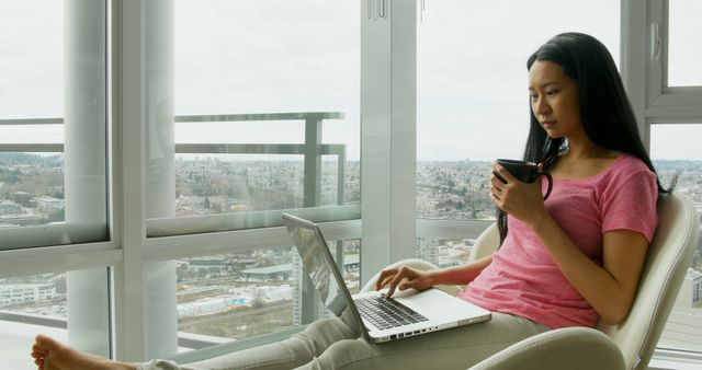 Young woman sitting in comfortable chair, working on laptop and holding coffee mug, in bright modern apartment with large windows and cityscape in background. Ideal for illustrations on work-from-home, relaxation, modern living, and technology use in everyday life settings.