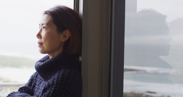 Woman standing by window, gazing outside and contemplating. Ideal for use in mental health awareness campaigns, articles on mindfulness, self-reflection, and relaxation techniques, or advertising casual wear and home environments promoting peaceful moments.
