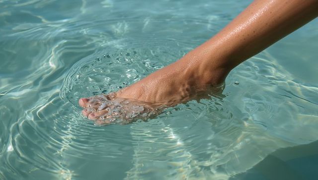 Sunlit foot entering turquoise pool water creating ripples, bubbles and caustics