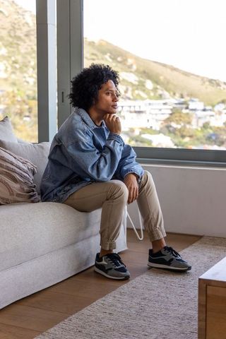 Pensive african american man sitting by window wearing denim jacket overlooking hillside