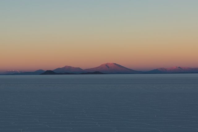 Serene and peaceful view of a mountain range illuminated by dawn light, seen over expansive salt flats. Ideal for promoting travel destinations, highlighting natural beauty, or illustrating concepts of tranquility and calm in nature.