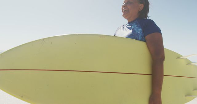 Woman in blue shirt smiling while holding a yellow surfboard on a sunny day at the beach. Great for themes of summer vacations, active outdoor lifestyles, surfing culture, sports, leisure, and nature enthusiasts.