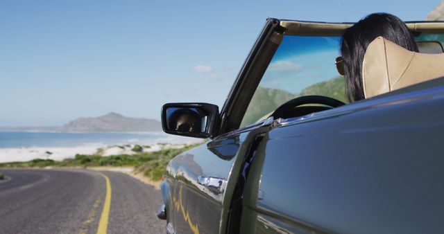 This image depicts a woman with long, dark hair wearing sunglasses driving a convertible car on a scenic coastal highway. The road curves left, with an ocean view and mountains in the background. Ideal for automotive advertising, travel blogs, tourism promotions, and lifestyle articles celebrating road trips, adventure, and the freedom of travel.