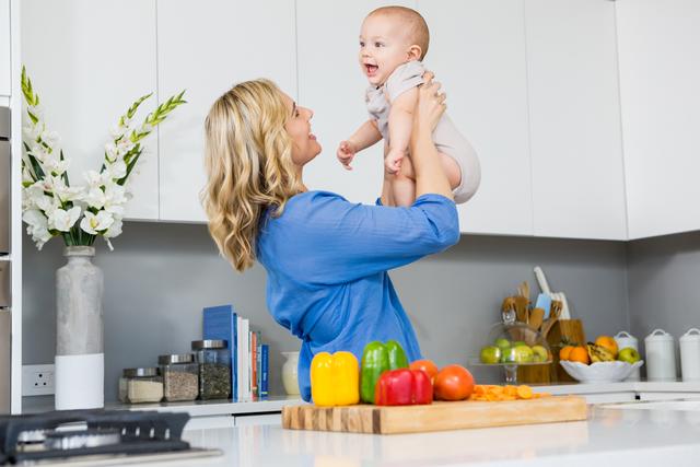 Mother holding her baby in a modern kitchen, both smiling and enjoying their time together. The kitchen counter has colorful bell peppers and other vegetables, suggesting a healthy lifestyle. Ideal for use in parenting blogs, family-oriented advertisements, and articles about healthy living and home life.
