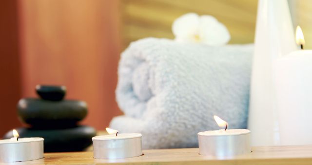 Lit candles provide a serene ambiance next to a soft towel and massage stones, suggesting a tranquil spa setting. The arrangement evokes a sense of relaxation and wellness, ideal for promoting self-care and therapeutic environments.